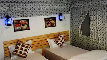 Quarto com duas camas de madeira, cada uma com colchão branco e almofada estampada. Há duas luminárias fixadas na parede, uma com luz azul e outra com luz branca. As paredes são decoradas com papel de parede floral, e há uma moldura com uma imagem na parede.