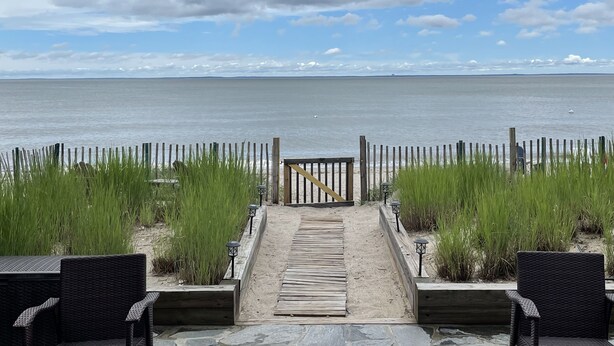 Ein Holzweg führt zu einem Strand mit Korbstühlen und einem Zaun.
