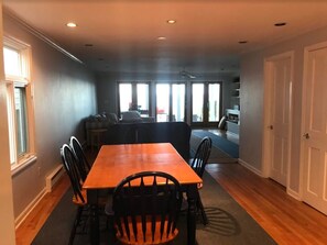 A dining area with a wooden table and chairs, a kitchen in the background, and a view through large windows. - Stunning direct waterfront beach house! (Fairfield)