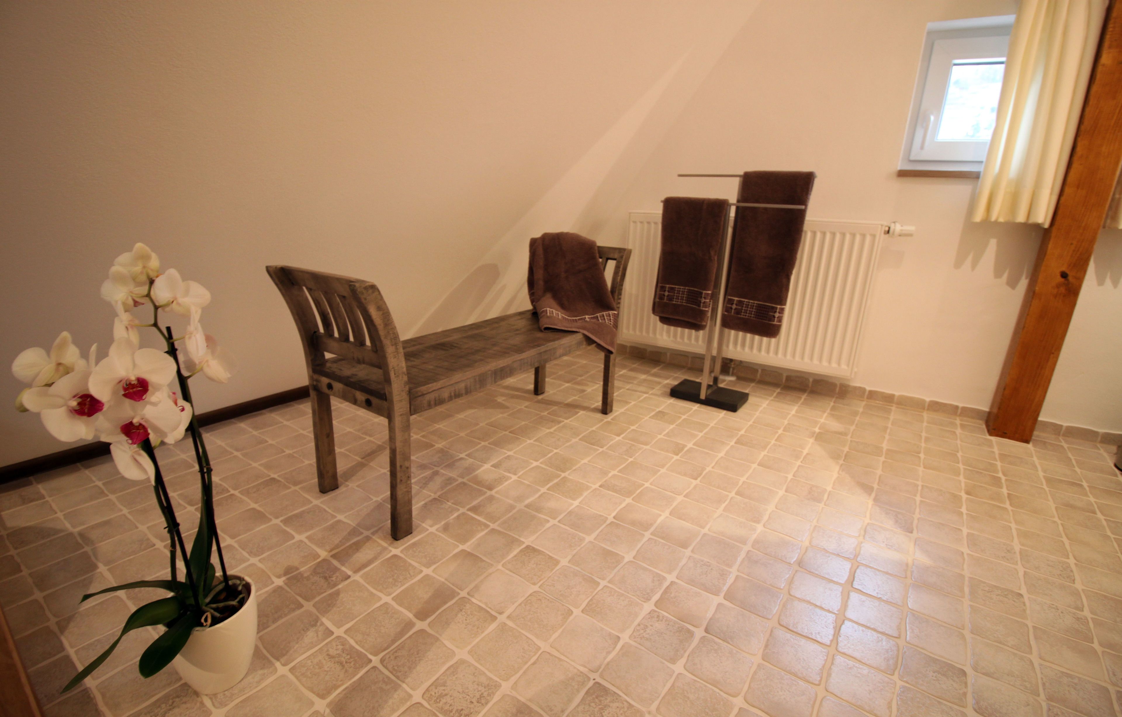Une pièce avec un sol carrelé, un banc en bois, une orchidée en pot et un radiateur sur lequel sont posées des serviettes.