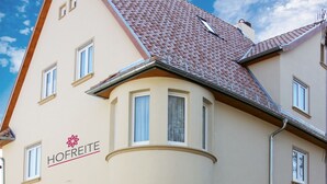 Un immeuble à plusieurs étages, doté d’une façade beige, d’un toit recouvert de tuiles rouges et de nombreuses fenêtres. L’immeuble arbore un logo représentant une fleur et le texte « HOFREIT ».