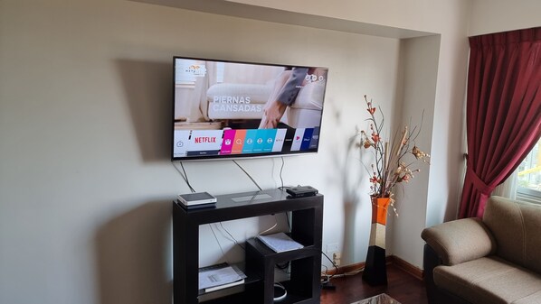 Un salon doté d’un téléviseur à écran plat fixé au mur, d’un meuble de rangement noir en dessous, d’un canapé beige et d’un vase décoratif orné de branches.