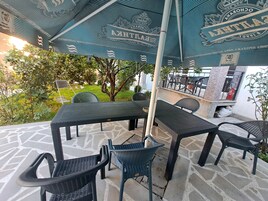 An outdoor patio area with a table and chairs under a blue umbrella, surrounded by greenery and a stone floor.