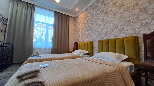 A hotel room with two beds, a large window with curtains, a radiator, and a wall with a patterned design. - Guest House Akhaltsikhe (Akhaltsikhe)