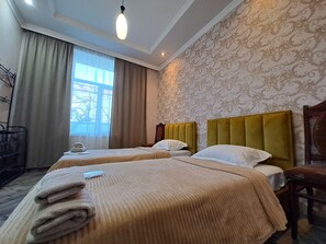 Ein Hotelzimmer mit zwei Betten, einem großen Fenster mit Vorhängen, einem Heizkörper und einer Wand mit einem gemusterten Design.
