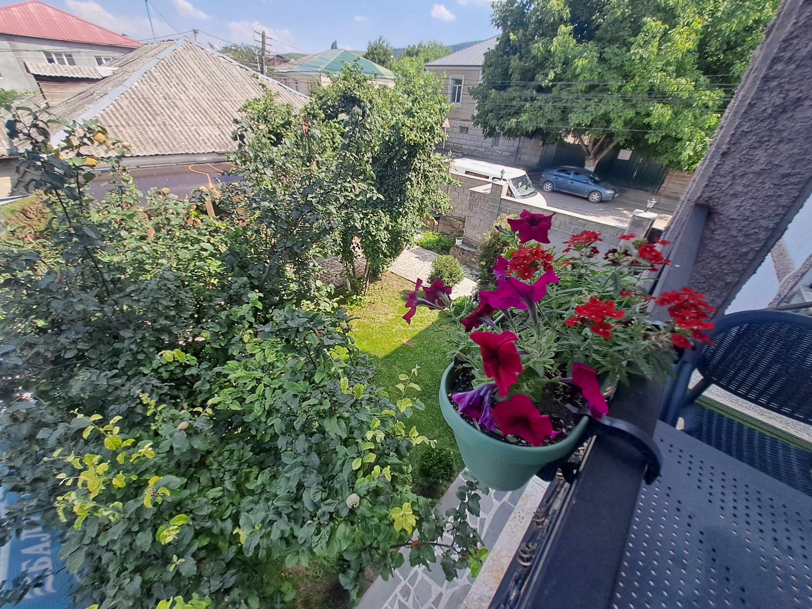 A balcony with a view of a garden, a parked car, and a building with a red roof.