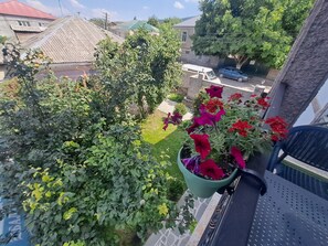 A balcony with a view of a garden, a parked car, and a building with a red roof. - Guest House Akhaltsikhe (Akhaltsikhe)