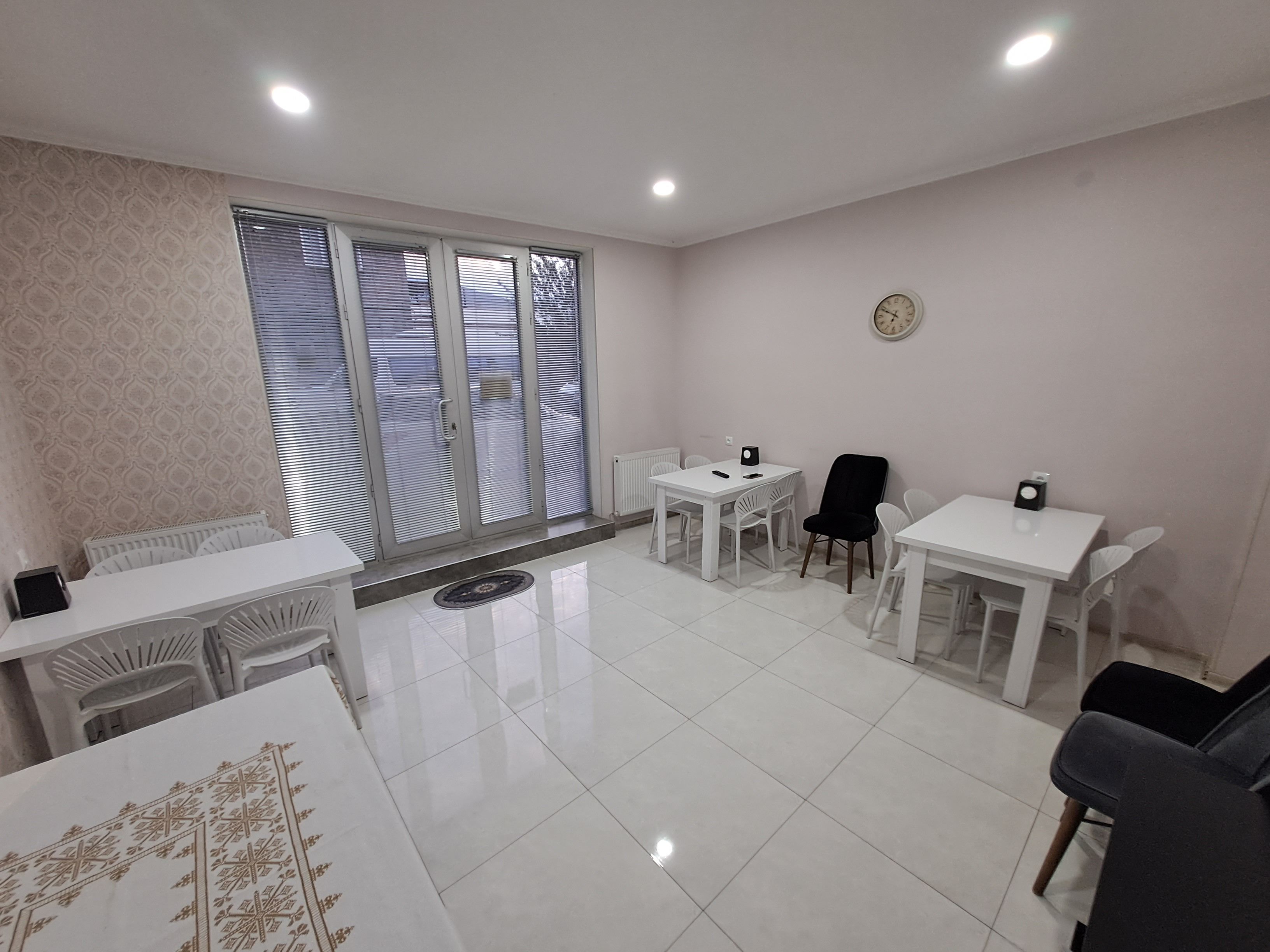 Una habitación moderna con piso de baldosas blancas, una mesa blanca con sillas, una silla negra, un radiador y una ventana con persianas.