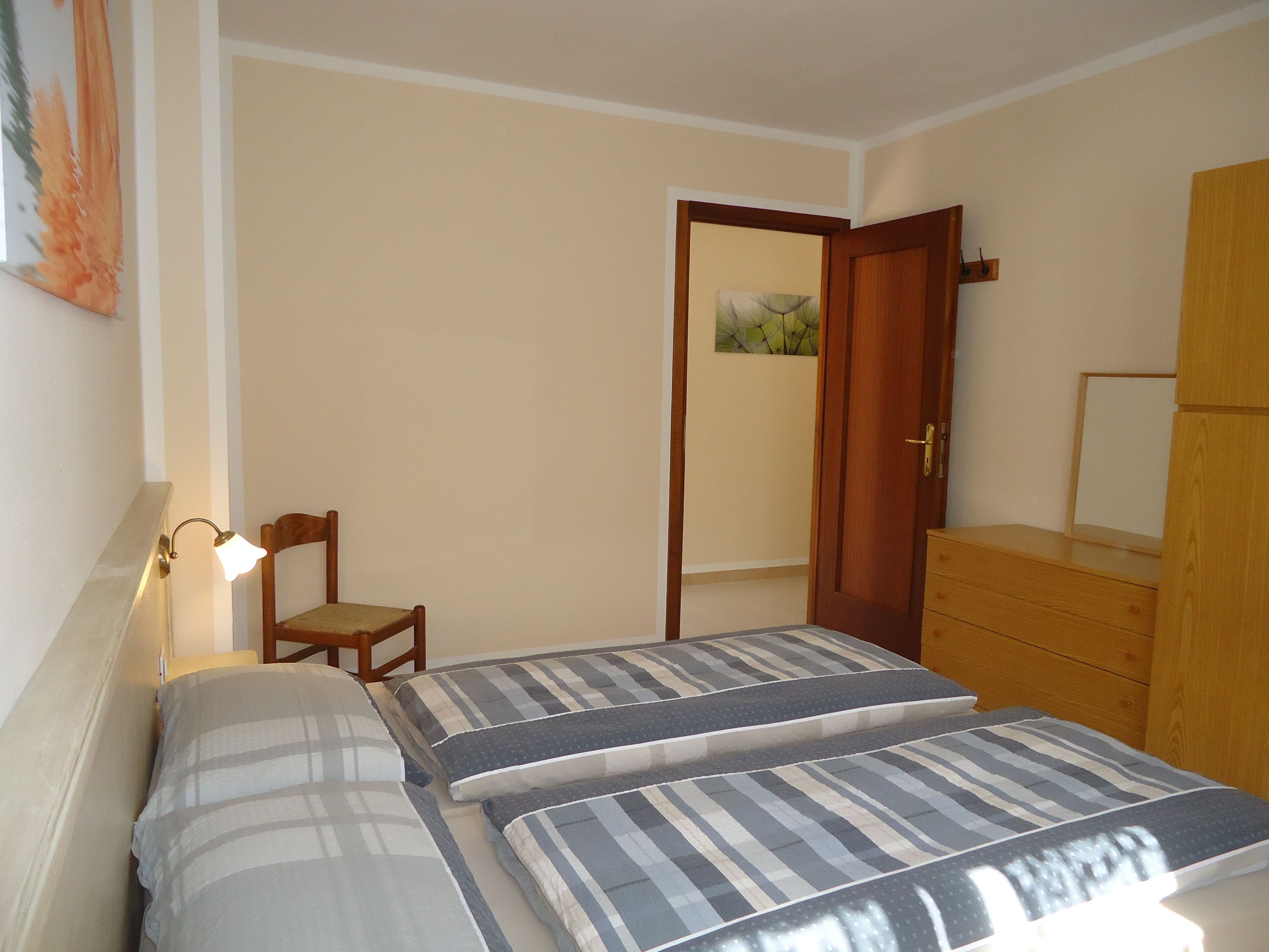 A bedroom with two beds, a chair, a wooden dresser, and a door leading to another room.