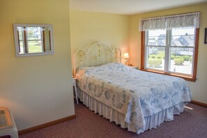 Quarto com cama, mesa de cabeceira, abajur, janela com vista para casas e uma pequena mesa com uma cadeira.