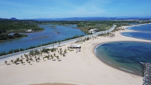 An aerial view of a coastal area with a beach, a river, and a road with palm trees.