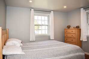 A bedroom with a bed, a window with curtains, a wooden dresser, and a headboard.