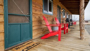 Een houten hut met veranda, rode Adirondack stoelen, een welkomstmat en een barbecue.