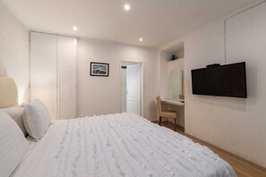 A bedroom with a bed, a chair, a desk with a mirror, and a TV.