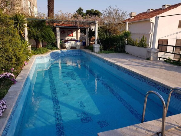 A swimming pool with a tiled edge, surrounded by greenery and a small structure with chairs.