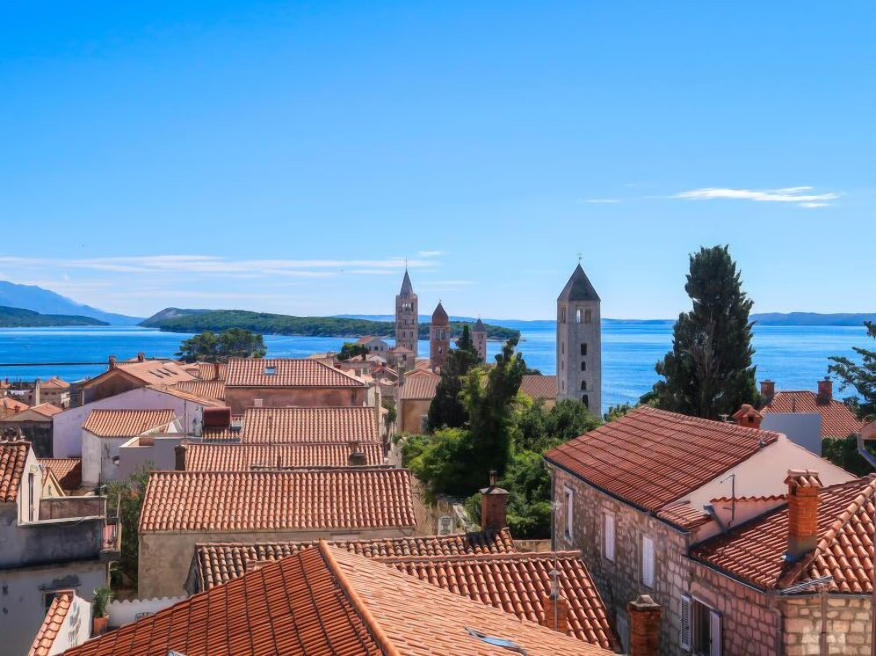 A coastal town with terracotta-roofed buildings, a prominent church with two towers, and a clear view of the sea.