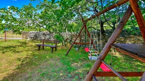 A wooden swing set in a grassy area with a stone wall and a bench in the background.