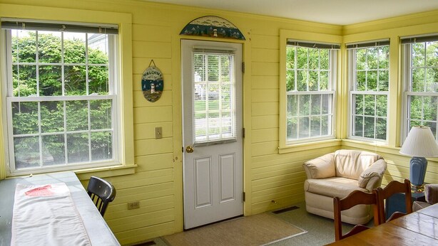 A room with yellow walls, a white door, and large windows. There is a beige chair and a table with a lamp.