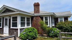 A two-story house with a brick chimney, white trim, and a small garden with purple flowers.
