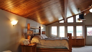 A bedroom with a wooden ceiling, a bed with a headboard, bedside lamps, a dresser, and a window with a view.