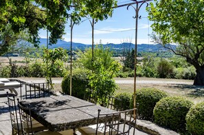 An outdoor dining area with a stone table and chairs, surrounded by greenery and a view of distant hills. - Fontblanche, Provencal estate for happy reunions (Gordes)