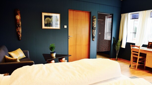 A bedroom with a large bed, a desk, and a chair. There is a door, a plant, and a framed picture on the wall. - Eco Camp Norway - Hostel (Smøla)
