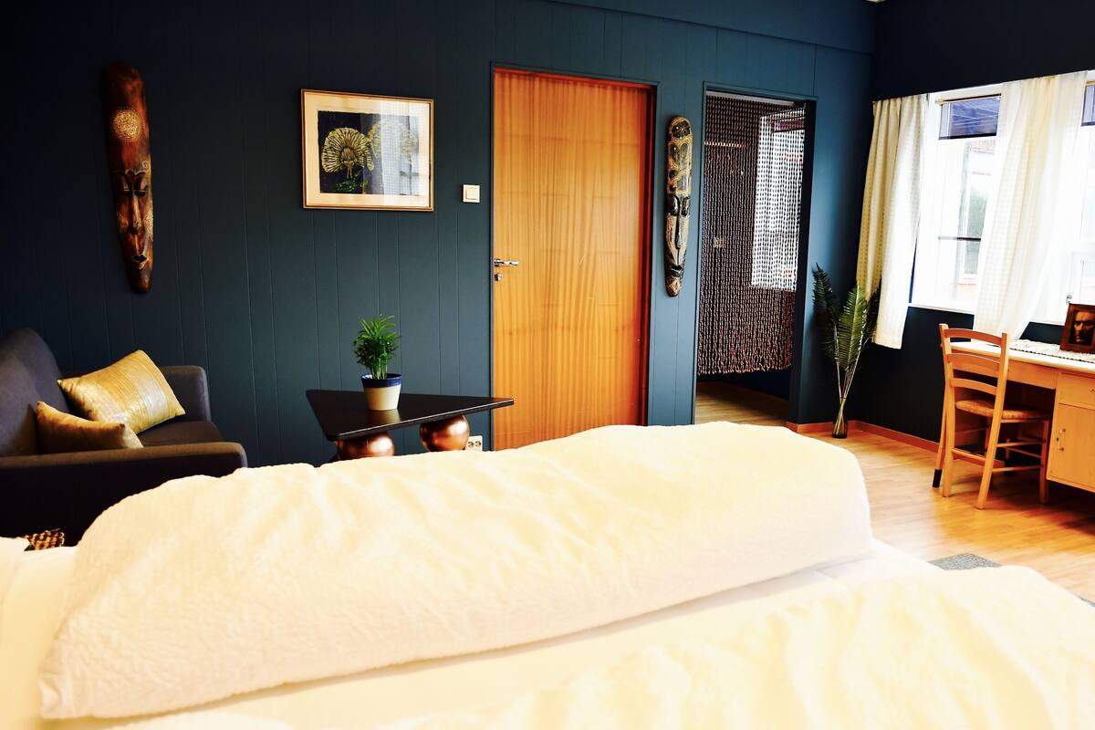 A bedroom with a large bed, a desk, and a chair. There is a door, a plant, and a framed picture on the wall.