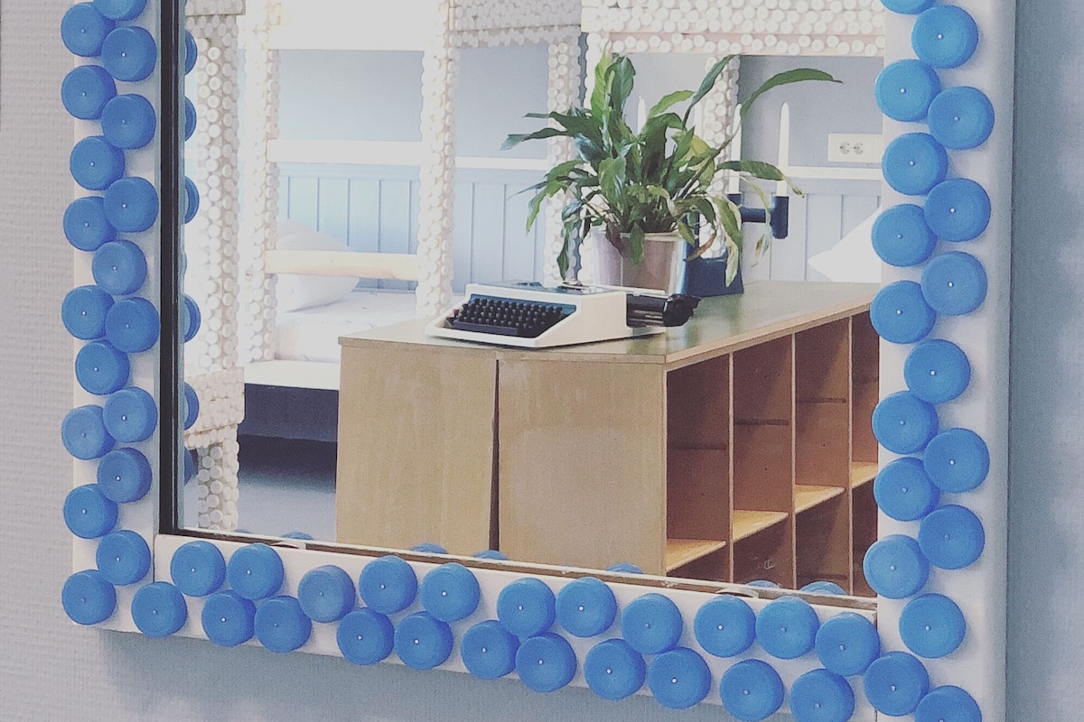 A mirror with a blue circular frame, reflecting a room with a white cabinet, a potted plant, and a typewriter.