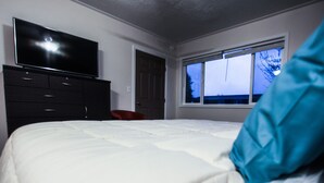 Un dormitorio con una cama, un televisor montado en una cómoda, una ventana con vista al cielo y una puerta.