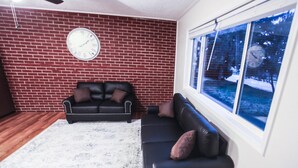 Un salón con pared de ladrillo, dos sofás negros, una alfombra blanca y un ventanal con vistas a árboles y nieve.