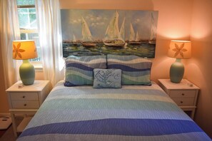 Ein Schlafzimmer mit einem Bett, zwei Nachttischen, zwei Lampen, einem Segelboot-Bild und einem Fenster mit Vorhängen.