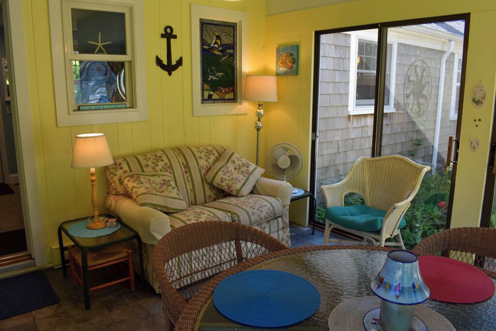 A cozy room with a floral-patterned sofa, wicker chairs, a small table with a lamp, and a sliding glass door leading to a garden.