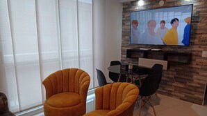 A modern living room with two mustard-colored armchairs, a glass coffee table, and a dining table with black chairs. A flat-screen TV mounted on a stone-patterned wall is displaying a group of people. - SUPERHOST APARTAMENTO, EXCELENTE UBICACION TULIPAN (Cúcuta)
