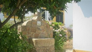 A stone structure with a plaque, surrounded by greenery and a building with a yellow wall and blue shutters. - Tapada da Beirã - Rural house (2-3 persons) (Marvão)