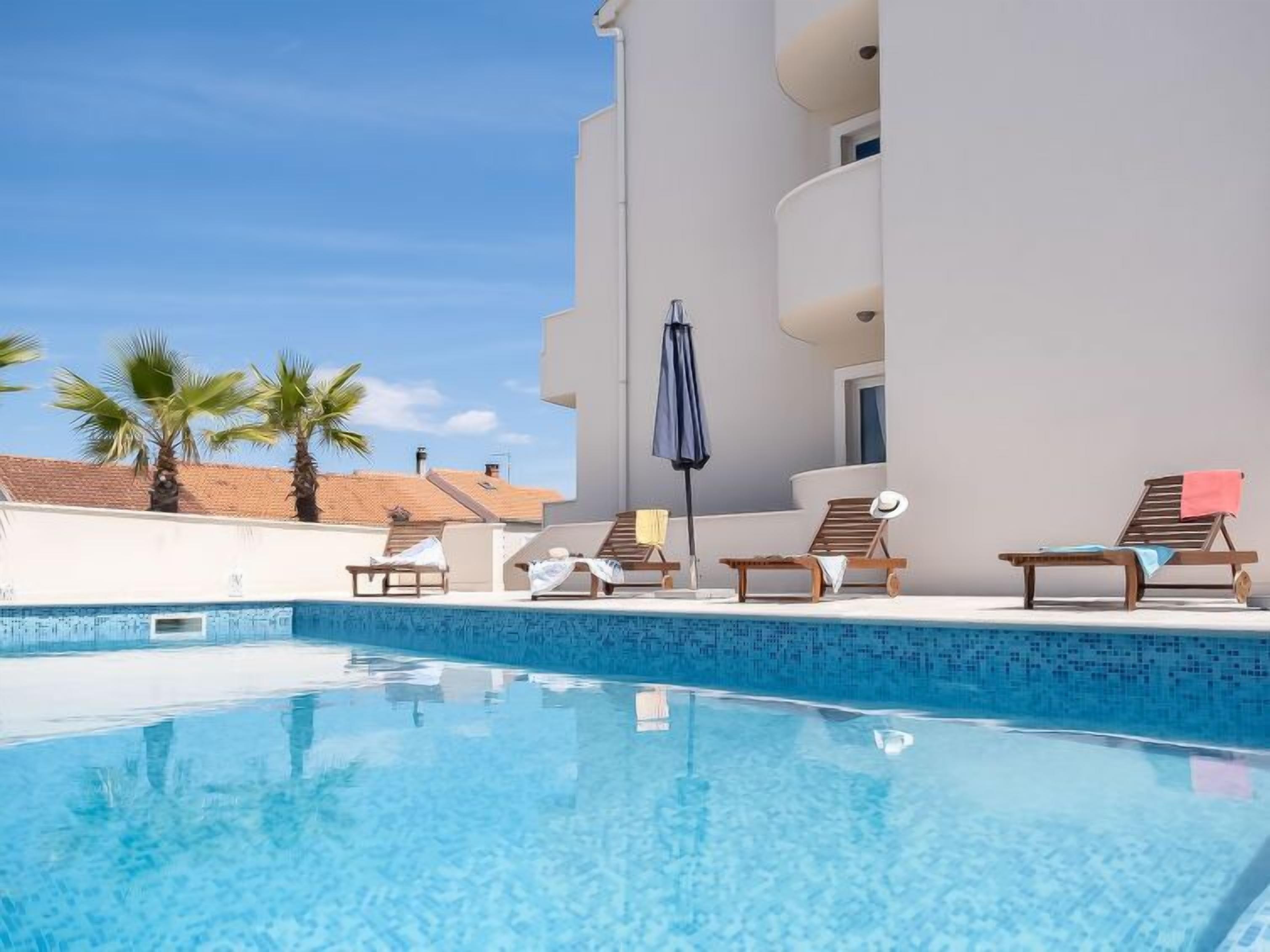 A swimming pool with lounge chairs and umbrellas, surrounded by a white building with balconies.