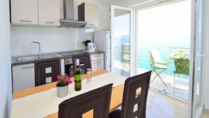 Une cuisine moderne avec des armoires blanches, un évier en acier inoxydable et un coin repas comprenant une table et des chaises. Un balcon avec vue sur la mer est visible à travers la porte ouverte.