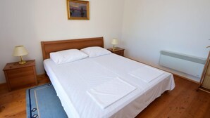 En pænt redt seng med hvide sengetøj, to sengeborde med lamper, et billede på væggen og en radiator.
