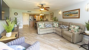 A living room with a sectional sofa, a flat-screen TV, a ceiling fan, and a kitchen in the background.