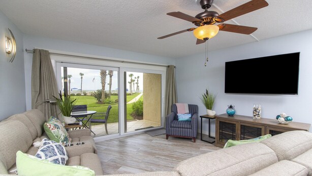 A spacious living room with a ceiling fan, a flat-screen TV, and a sliding glass door leading to a patio with a view of the ocean.
