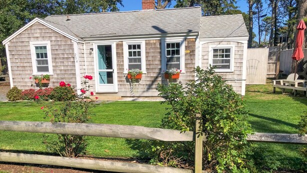A single-story house with a front door, two windows, and a small porch with potted plants. - Three bedroom cottage-Only .2 miles to Sea Street Beach (Dennis Port)