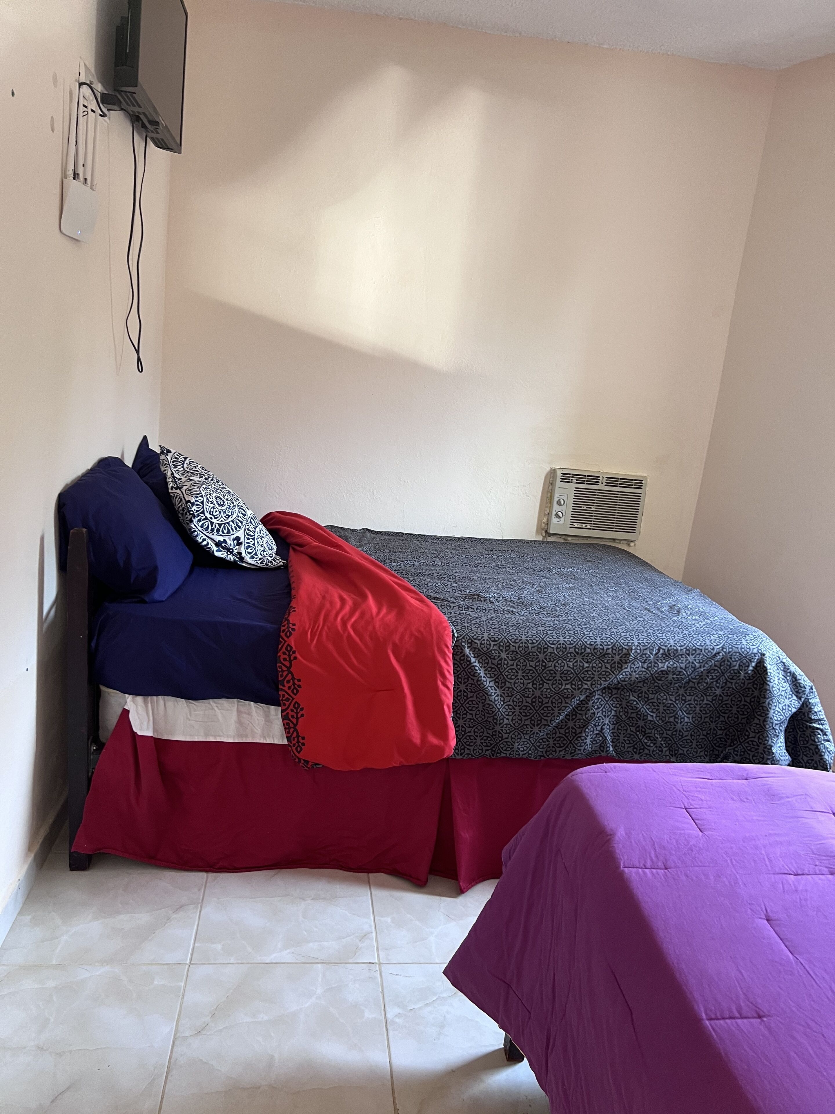 A small, single-bed room with a plain white wall, a window, and a wall-mounted air conditioning unit.