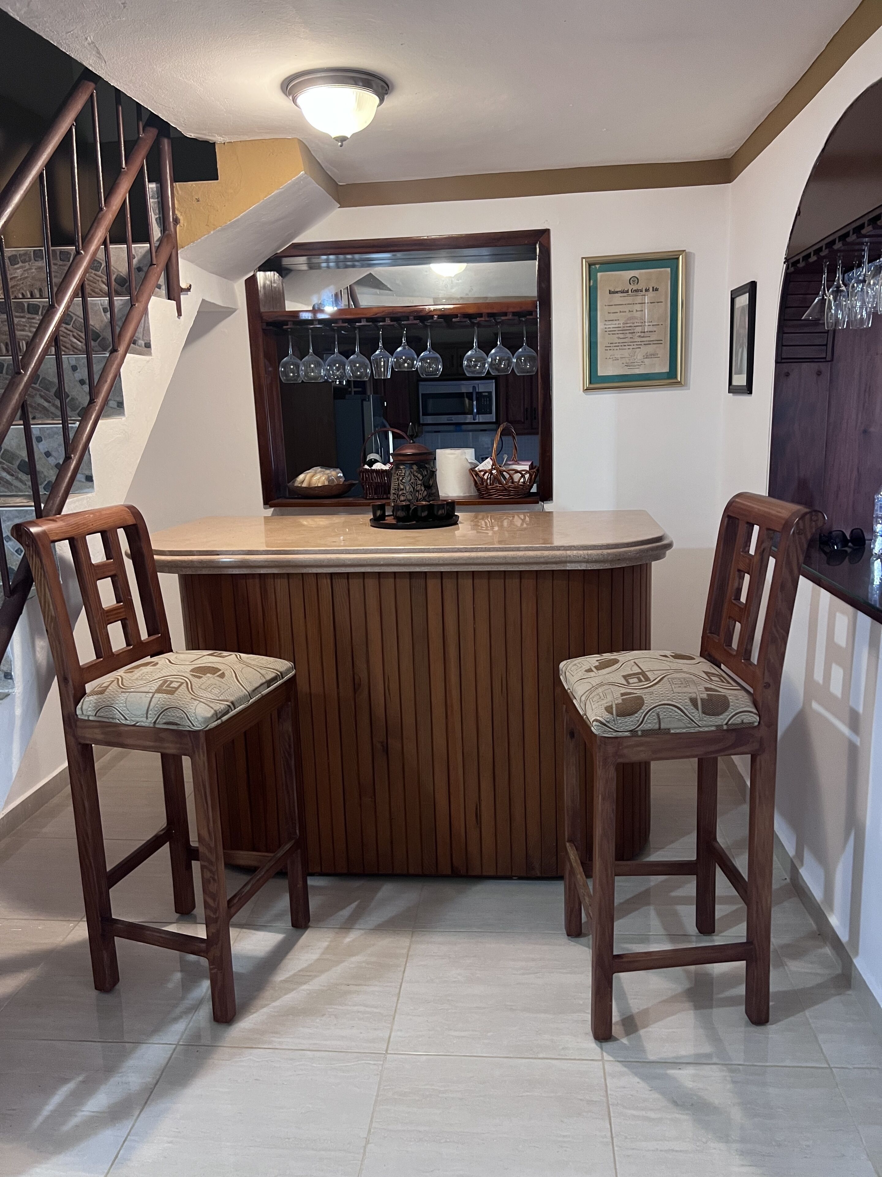 A kitchen area with a wooden bar counter, two wooden bar stools, a microwave, and hanging wine glasses.