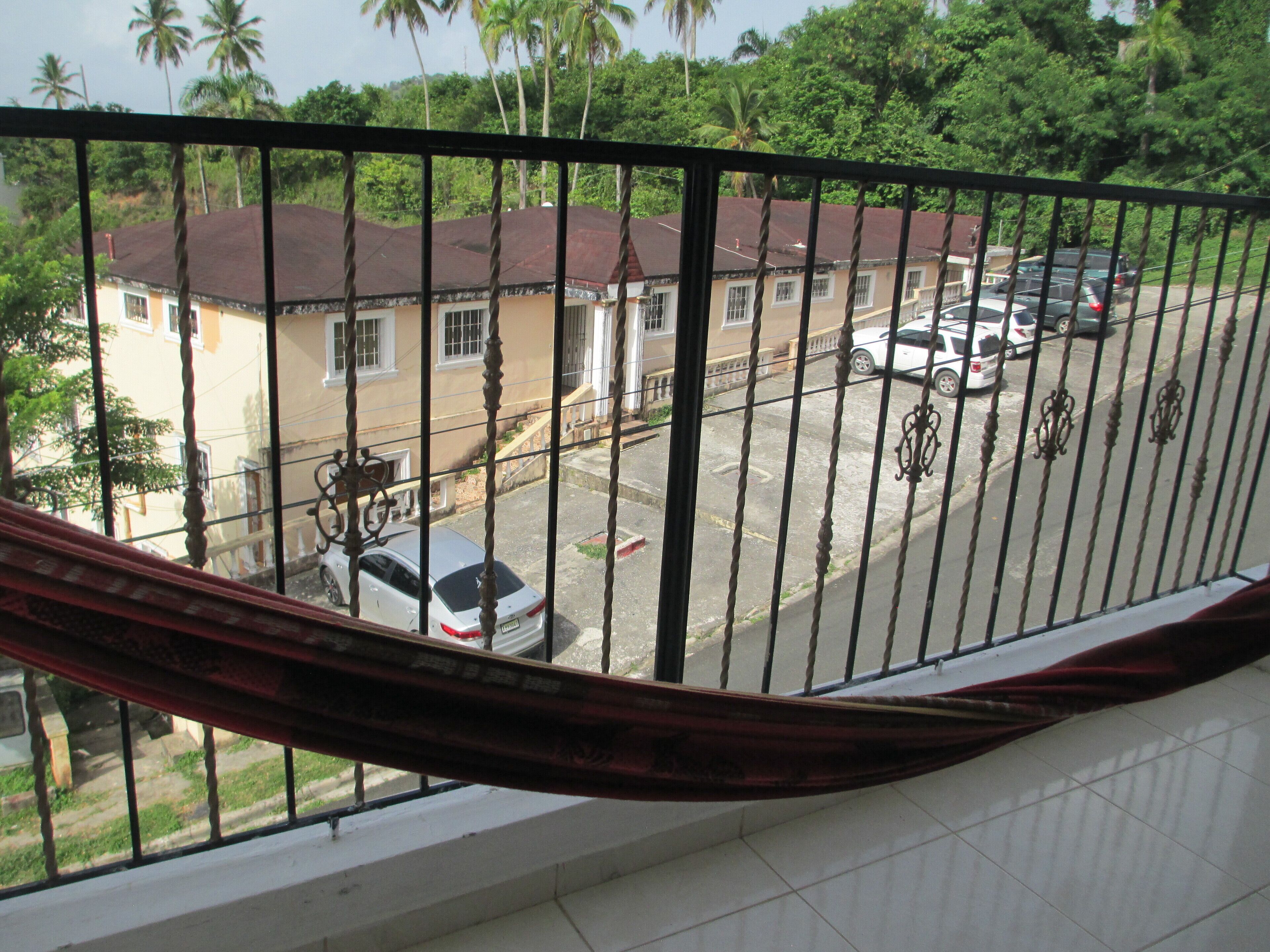 A view from a balcony with a metal railing, overlooking a street with parked cars and a building with a brown roof.