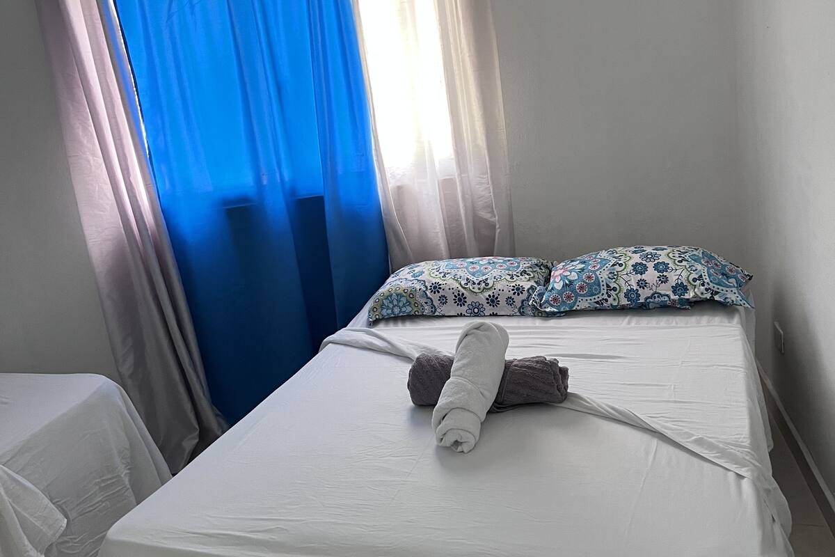 Hotellihuone, jossa on sänky, rullalle taitettu pyyhe ja kukkakuviollinen tyyny.