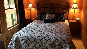 A bed with a patterned bedspread, two bedside lamps, and a window with a view of greenery. - Little Beans Cottage on Great Pond, Belgrade ME (Rome)