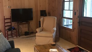 A cozy cabin interior with a wooden floor, a striped armchair, a television, and a wooden coffee table. - Little Beans Cottage on Great Pond, Belgrade ME (Rome)
