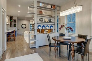 Un espace repas moderne avec une table ronde, des chaises et un lustre. On y trouve également un coin cuisine avec un plan de travail blanc et une horloge au mur.