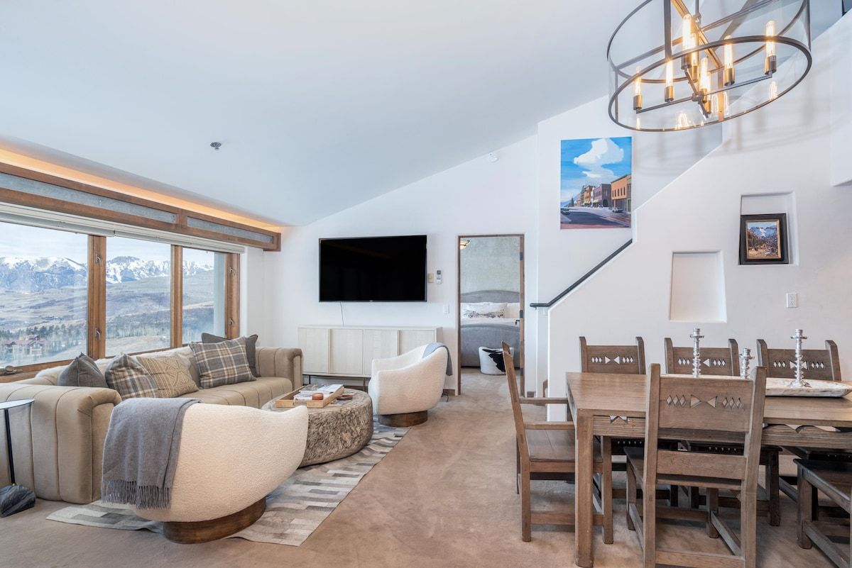 A modern living room with a sofa, armchairs, a dining table with chairs, a flat-screen TV mounted on the wall, and a staircase leading to another level.