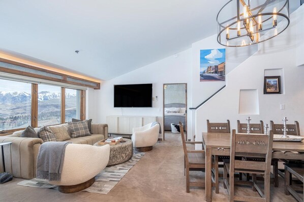 A modern living room with a sofa, armchairs, a dining table with chairs, a flat-screen TV mounted on the wall, and a staircase leading to another level.