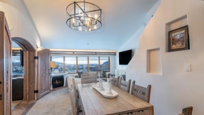 Comedor con mesa de madera, sillas y candelabro. Cuenta con chimenea y vistas a las montañas a través de amplias ventanas.